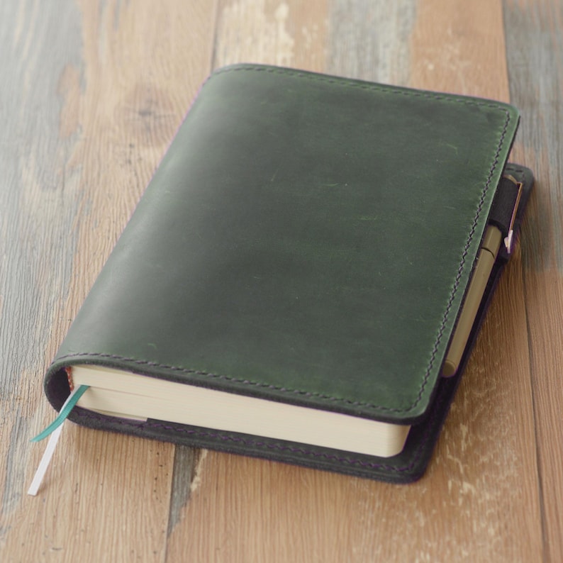 May include: A dark green leather journal with a pen holder and a light blue and white ribbon bookmark. The journal is closed and resting on a wooden surface. The cover is stitched with dark green thread.