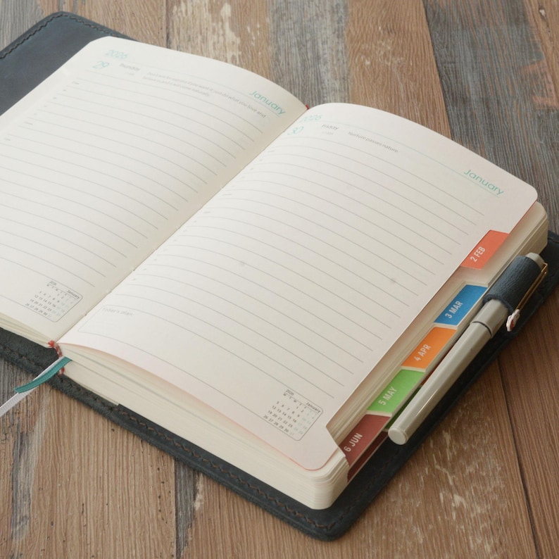 May include: An open, lined journal with colorful month tabs and a pen. The journal is black with a leather-like texture. The tabs are orange, blue, green, and red. The pen is silver and black.