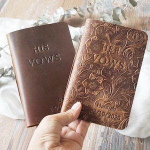 May include: Two leather vow books. One is embossed with "HIS VOWS" and the other with "HER VOWS". The "HER VOWS" book has a floral pattern and the name "Hannah" and date "08/10/2026" embossed on it. The "HIS VOWS" book has the date "08/17/" embossed on it.