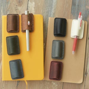 Könnte beinhalten: Eine Sammlung rechteckiger Stiftehalter aus Leder in verschiedenen Farben wie Braun, Schwarz, Grün, Creme und Burgunderrot. Diese Halter sind an einem gelben und einem beigen Notizbuch befestigt, wobei zwei Stifte in die Halter gesteckt sind.