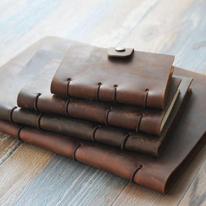 May include: A stack of brown leather journals in various sizes. The journals have a rustic, worn appearance, with visible stitching along the spine and a button closure on the smallest journal. The leather has a rich, warm tone, suggesting a handmade or vintage aesthetic.