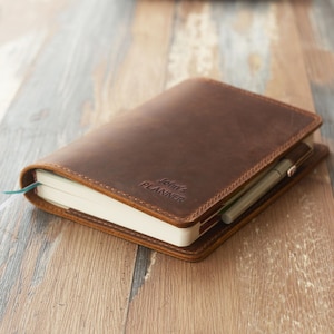 May include: A brown leather planner with the text "John's PLANNER" embossed on the cover. The planner is open, revealing white pages and a pen tucked inside. A blue ribbon bookmark is visible.