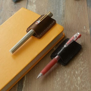 Könnte beinhalten: Ein dunkelbrauner Stiftehalter aus Leder, der an einem gelben Notizbuch befestigt ist, mit weißen Nähten und einem kleinen Hasensymbol am Stifteclip. Daneben hält ein schwarzer Lederhalter einen roten Gelschreiber mit transparenter Kappe und der Aufschrift Pilot Juice.