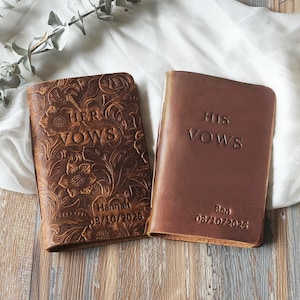 May include: Two leather vow books. One is embossed with floral patterns and the words "HER VOWS", the name "Hannah", and the date "08/10/2026". The other is a solid brown with "HIS VOWS", the name "Ben", and the date "08/10/2026".