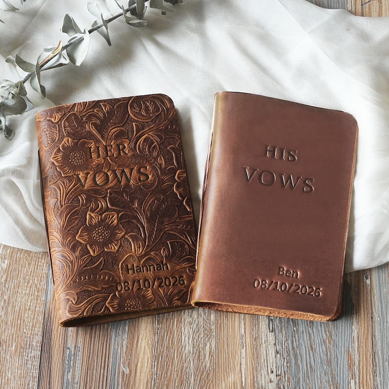May include: Two leather vow books. One is embossed with floral patterns and the words "HER VOWS", the name "Hannah", and the date "08/10/2026". The other is a solid brown with "HIS VOWS", the name "Ben", and the date "08/10/2026".