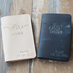 May include: Two leather-bound vow books, one white and one black, with the words "Her Vows" and "His Vows" in cursive script. The books are personalized with names and dates: "Maddy 08.10.2023" and "Logan 08.10.2023".