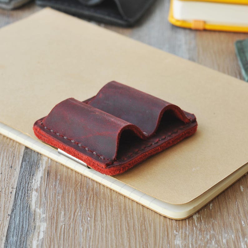 May include: A burgundy leather pen holder with two slots. The holder is stitched with a contrasting thread and has a slightly wavy design.