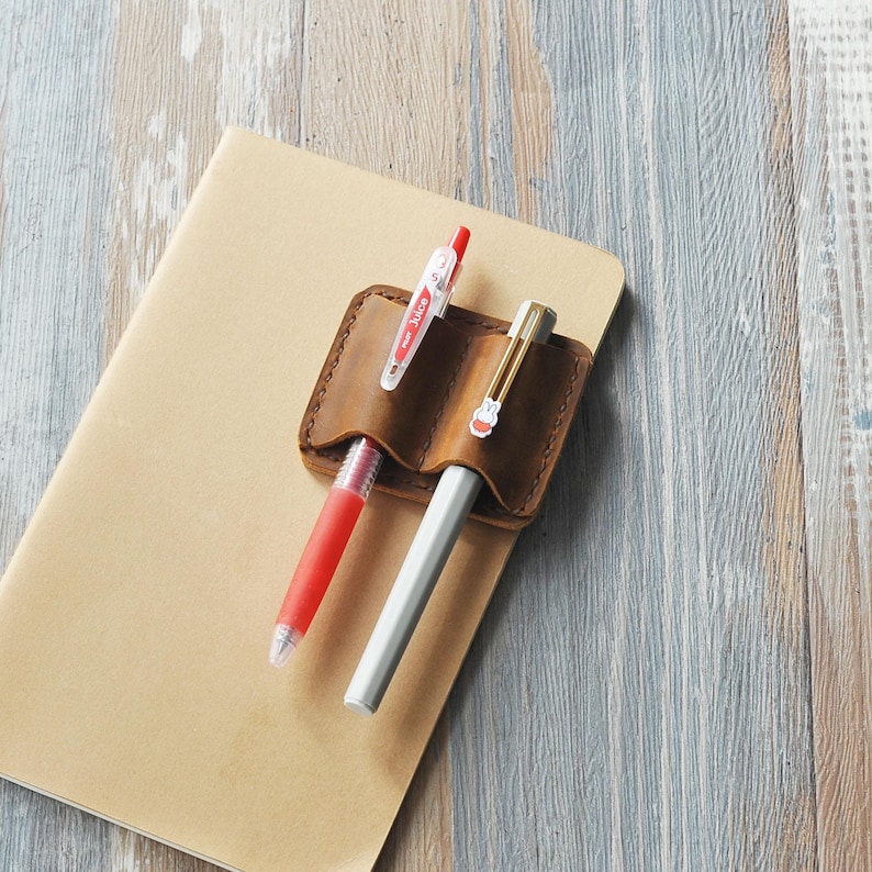 May include: A brown leather pen holder with two slots for pens. A red and clear pen and a silver pen are inserted in the slots. The pen holder is on top of a brown notebook.