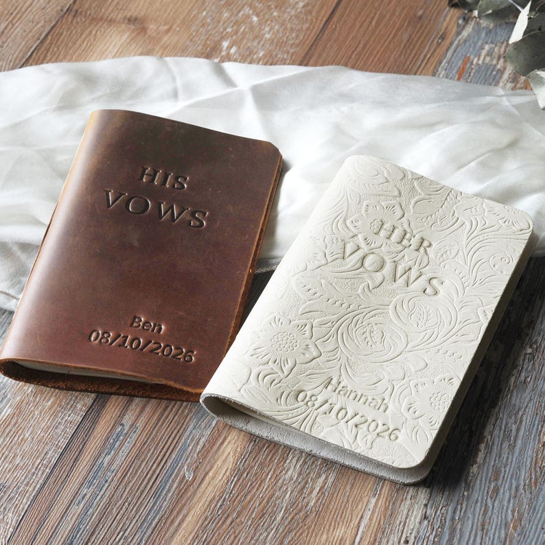 May include: Two leather vow books. One is brown with "HIS VOWS" embossed on the cover, along with the name "Ben" and the date "08/10/2026". The other is white with "HER VOWS" and floral detailing, with the name "Hannah" and the date "08/10/2026".