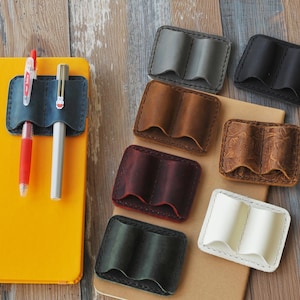 May include: Assortment of leather pen holders in various colors and styles. The holders are made of genuine leather and have two slots for pens. The holders are displayed on a wooden surface.