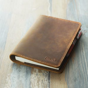 May include: A brown leather journal with the embossed word "Emmit" on the cover. The journal is closed, with a pen visible in the spine. The leather has a distressed appearance, and the edges are stitched.
