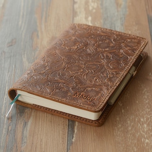 Op de afbeelding: Een bruin leren notitieboek met een bloemreliëf. Het woord "Maddy" is in de omslag geëmbosseerd. Een pen zit in het notitieboek, met een groene lint-bladwijzer.