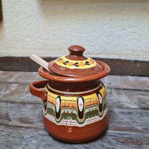 Puede incluir: Una pequeña olla de cerámica marrón con tapa y cuchara de madera. La olla tiene un asa y está decorada con patrones coloridos en amarillo, naranja, verde y blanco. La tapa está adornada con un diseño floral.