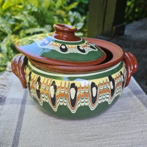 Puede incluir: Una olla de cerámica verde y marrón con tapa. La olla tiene un patrón decorativo de remolinos negros, blancos y naranjas. La tapa tiene un pomo marrón.