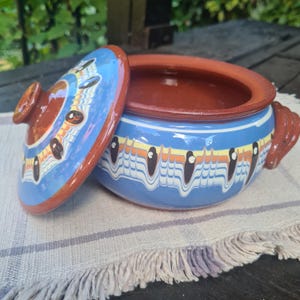 Pode incluir: Uma panela redonda de terracota com tampa a condizer, apresentando um exterior vidrado em azul claro decorado com um padrão repetitivo de motivos em forma de gota em castanho escuro, amarelo e branco. O interior e o rebordo são de argila natural.