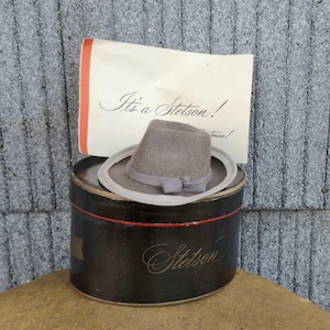May include: A gray Stetson fedora hat with a light gray brim and a matching ribbon band, resting on a black hat box with the name "Stetson" in elegant script. A card with the text "It's a Stetson!" is behind the hat.