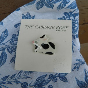 May include: A ceramic bunny pin with black spots and pink accents, resting on a card that reads "THE CABBAGE ROSE" by Patti Bax. The pin is set against a blue and white floral patterned fabric.