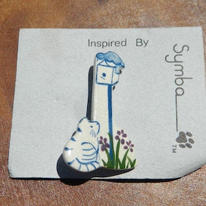 May include: A ceramic pin featuring a whimsical design of a birdhouse, a cat, and flowers. The pin is primarily white with blue accents and touches of green and purple. The pin is on a gray card with the text "Inspired By" and the brand name "Symba".