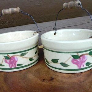 May include: Two cream-colored ceramic buckets with wire handles and wooden grips. Each bucket has a green band and a floral design with pink flowers and green leaves. The buckets are displayed on a wooden surface.