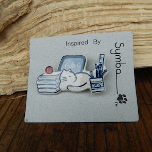 May include: A ceramic pin featuring a white cat resting on a stack of books, with a laptop and art supplies. The pin is on a gray card with the text "Inspired By Symba". A pink ball sits on top of the books.
