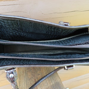 Puede incluir: Bolso de mano de cuero marr&oacute;n con m&uacute;ltiples compartimentos. El interior presenta un forro oscuro estampado y una tela a rayas. El bolso tiene un asa trenzada y herrajes plateados. El bolso est&aacute; abierto, revelando el interior.