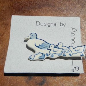 May include: A ceramic pin featuring a mother duck and her ducklings, in white and blue. The pin is on a light gray card with the text "Designs by Anna". The pin is a charming accessory.