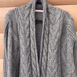 Puede incluir: Un c&aacute;rdigan gris de punto trenzado, abierto por delante, con cuello chal. El su&eacute;ter est&aacute; colgado de una percha negra sobre un fondo de madera marr&oacute;n claro. El patr&oacute;n de punto trenzado es visible en todo el su&eacute;ter.