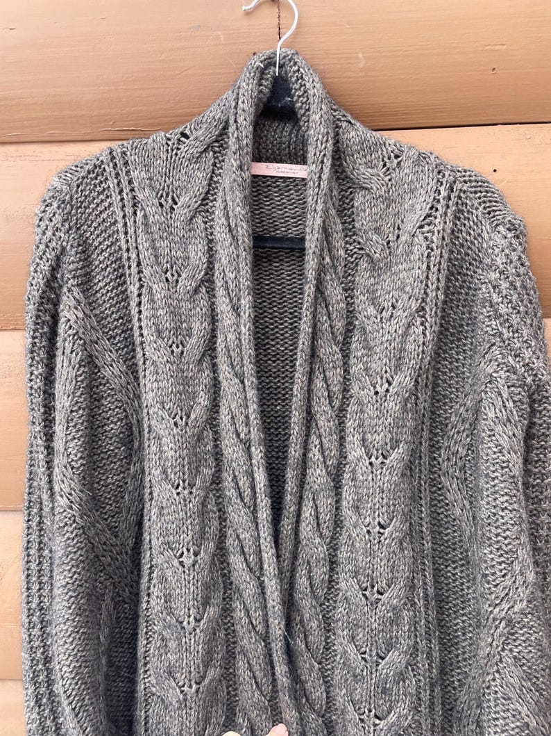Puede incluir: Un c&aacute;rdigan gris de punto trenzado, abierto por delante, con cuello chal. El su&eacute;ter est&aacute; colgado de una percha negra sobre un fondo de madera marr&oacute;n claro. El patr&oacute;n de punto trenzado es visible en todo el su&eacute;ter.