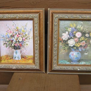 May include: Two framed floral paintings. The paintings depict bouquets of colorful flowers in vases. The frames are gold-toned with ornate detailing. The background is a wooden surface.