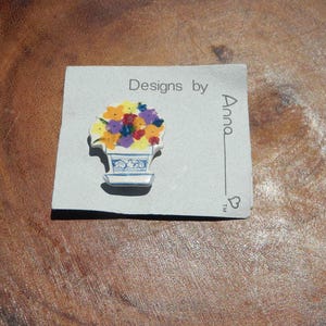 May include: A ceramic pin featuring a bouquet of colorful flowers in a blue and white vase. The pin is on a gray card with the text "Designs by Anna". The flowers are yellow, orange, red, and purple.