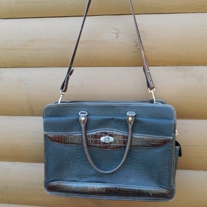 Puede incluir: Un malet&iacute;n negro con detalles marrones de imitaci&oacute;n de cocodrilo y un asa a juego. El bolso tiene una correa larga y ajustable para el hombro y un cierre plateado. El malet&iacute;n est&aacute; colgado sobre un fondo de madera.