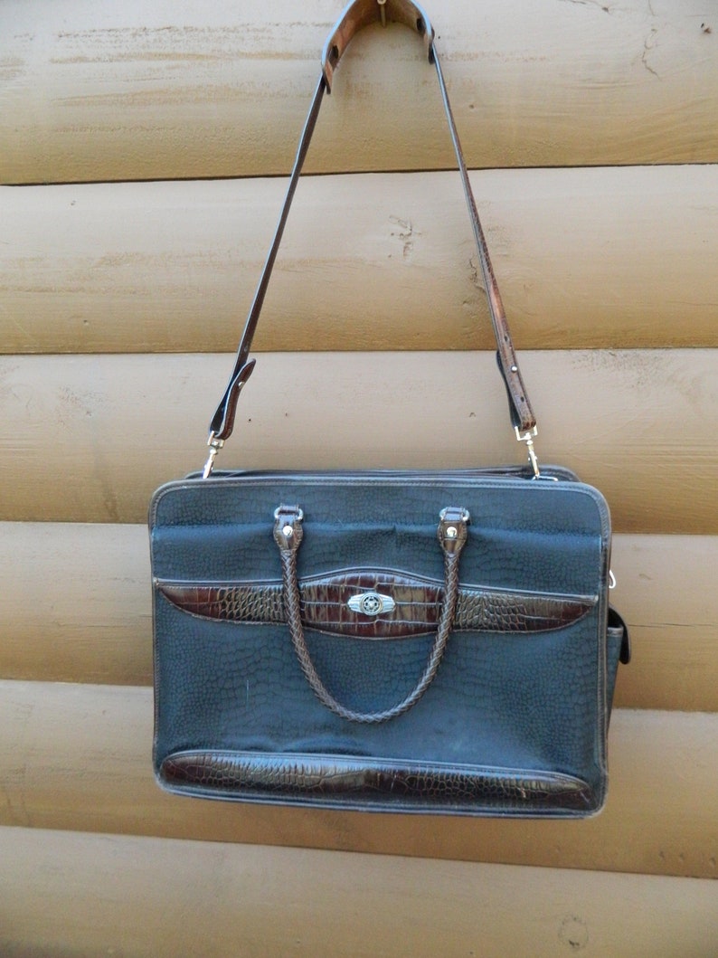 Puede incluir: Un malet&iacute;n negro con detalles marrones de imitaci&oacute;n de cocodrilo y un asa a juego. El bolso tiene una correa larga y ajustable para el hombro y un cierre plateado. El malet&iacute;n est&aacute; colgado sobre un fondo de madera.