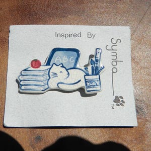 May include: A ceramic pin featuring a white cat resting on a stack of books, with a small red apple, a chalkboard with "ABC", and a pencil holder. The pin is blue and white, with the text "Inspired By" and "Symba".