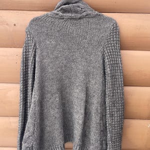 Puede incluir: Un su&eacute;ter de punto gris con cuello alto, colgado de una percha blanca. El su&eacute;ter presenta un patr&oacute;n de punto texturizado en el cuerpo y las mangas, con detalles de punto trenzado en los laterales. La prenda est&aacute; hecha de un material suave y c&aacute;lido.