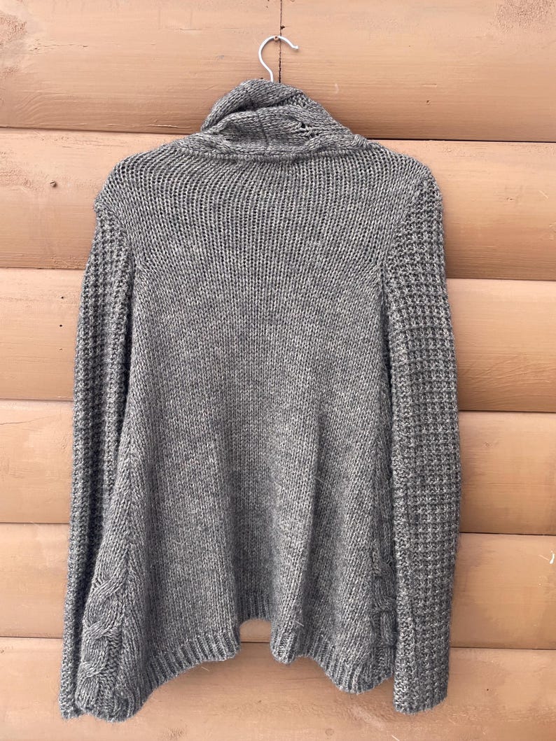 Puede incluir: Un su&eacute;ter de punto gris con cuello alto, colgado de una percha blanca. El su&eacute;ter presenta un patr&oacute;n de punto texturizado en el cuerpo y las mangas, con detalles de punto trenzado en los laterales. La prenda est&aacute; hecha de un material suave y c&aacute;lido.