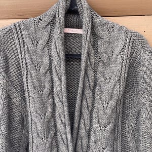 Puede incluir: Un c&aacute;rdigan gris tejido con trenzas y cuello chal. El su&eacute;ter est&aacute; hecho de un hilo grueso y texturizado y presenta intrincados patrones de trenzas en la parte delantera y en las mangas. El c&aacute;rdigan est&aacute; colgado en una percha blanca.
