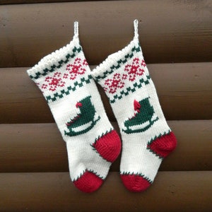 May include: Two knitted Christmas stockings. The stockings are white with red and green accents. Each stocking has a red heel and toe, a green ice skate design, and a red and green pattern at the top. They hang against a brown wooden background.