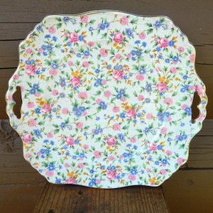 May include: A decorative serving tray with a floral pattern. The tray has a scalloped edge and two handles. The floral pattern features pink roses, blue flowers, and yellow accents on a cream background. The tray has a gold trim.