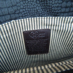 Puede incluir: Vista interior de un bolso de mano azul oscuro texturizado. El forro presenta un patr&oacute;n de rayas crema y azul. Una etiqueta de cuero marr&oacute;n oscuro con el nombre de la marca y texto es visible.