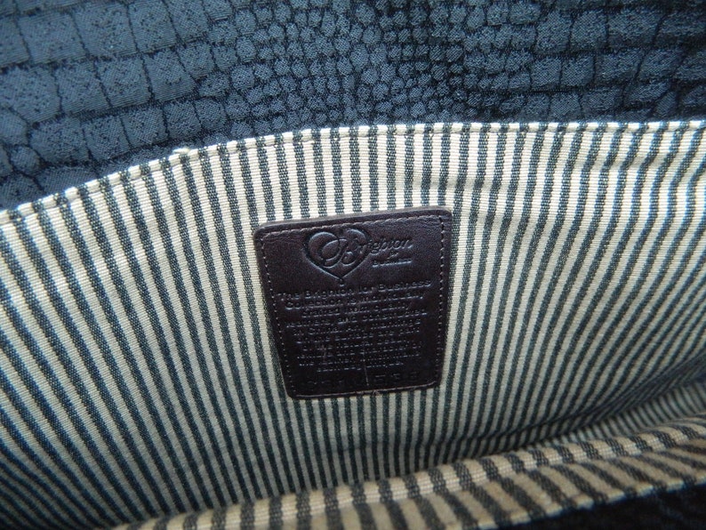 Puede incluir: Vista interior de un bolso de mano azul oscuro texturizado. El forro presenta un patr&oacute;n de rayas crema y azul. Una etiqueta de cuero marr&oacute;n oscuro con el nombre de la marca y texto es visible.