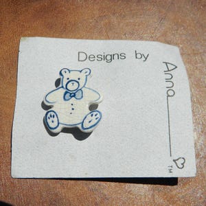 May include: A white ceramic teddy bear pin with blue accents, including a bow tie and paw details. The pin is on a card that reads "Designs by Anna". The bear has a cracked glaze effect.