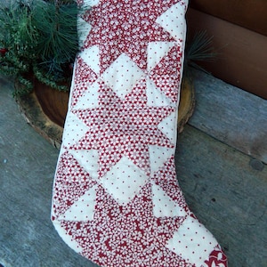 May include: A quilted Christmas stocking featuring a repeating star pattern in deep red and cream. The fabric displays a mix of small floral prints and tiny polka dots, with a white fabric loop attached to the top corner for hanging purposes.
