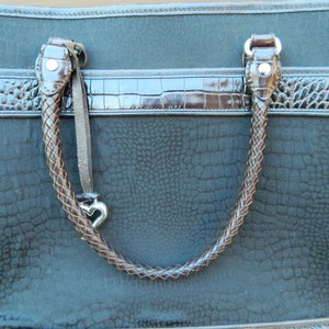 Puede incluir: Bolso tote de color verde azulado oscuro con asas trenzadas marrones y ribete marr&oacute;n de imitaci&oacute;n de cocodrilo. El bolso presenta un patr&oacute;n texturizado y un dije plateado en forma de coraz&oacute;n colgando del asa.