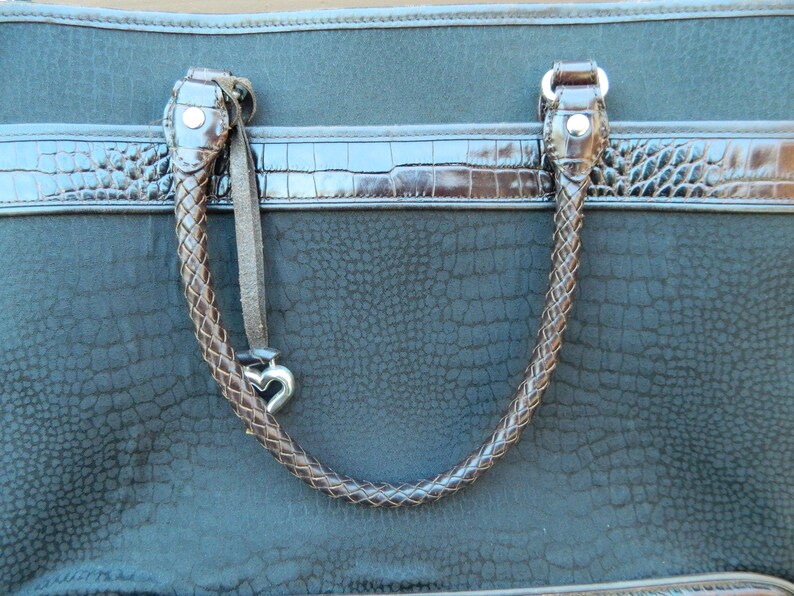 Puede incluir: Bolso tote de color verde azulado oscuro con asas trenzadas marrones y ribete marr&oacute;n de imitaci&oacute;n de cocodrilo. El bolso presenta un patr&oacute;n texturizado y un dije plateado en forma de coraz&oacute;n colgando del asa.