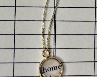 May include: A gold-colored necklace with a delicate chain and a round pendant. The pendant contains the word "home" in a clear, circular frame. The background features a grid pattern with the words "DATE DUE" and the date "APR 18 1996".