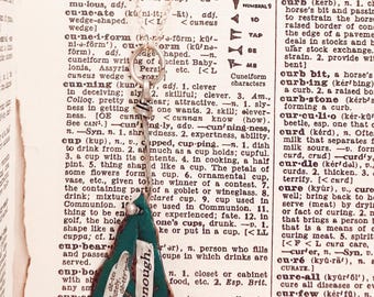 May include: A silver-colored necklace pendant in the shape of a leaf, with a green fabric insert. The leaf has the text "I was determined enough" printed on it. The pendant hangs against a backdrop of dictionary pages.