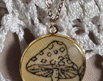 May include: A gold-toned circular pendant necklace featuring a black line-art illustration of a mushroom on a textured cream-colored background. The pendant is suspended from a matching gold-toned metal chain link necklace.