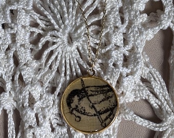 May include: A gold-toned circular pendant necklace featuring a tan fabric insert with a black line drawing of a bird. The pendant is suspended from a delicate gold-toned metal chain. The design is encased in a thin gold-toned metal frame.