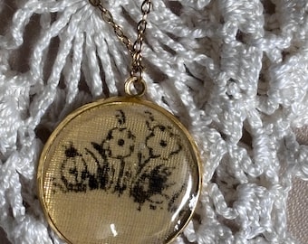 May include: A round gold-toned pendant necklace featuring a beige fabric insert with a black ink illustration of three flowers. The pendant is suspended from a delicate gold-toned metal chain, showcasing a clear, smooth, and slightly domed glass cover.