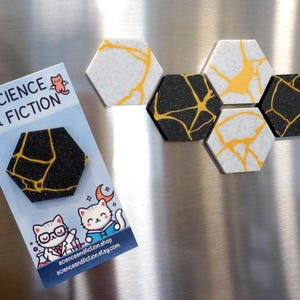 May include: A collection of hexagonal magnets with a Kintsugi design. Some are black with gold-colored lines, others are white with yellow lines. A card with the text "SCIENCE & FICTION" and cat illustrations is also present.